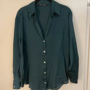 Mörkgrön skjorta från Zara - Snygg mörkgrön skjorta från Zara med klassisk krage och knappar framtill. Skjortan har lång ärm med bred manschett och är tillverkad i ett mjukt, lätt material som ger en skön känsla. Perfekt för dig som gillar stilrena plagg med enkel design.
