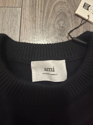 Svart stickad tröja från Ami Paris - Svart stickad tröja från Ami Paris med rund halsringning och ribbade muddar. På bröstet finns ett stort rött hjärta med bokstaven A, vilket ger en unik och trendig look. Perfekt för dig som gillar stilrena plagg med en twist.