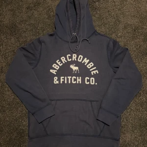 Mörkblå hoodie Abercrombie & Fitch - Mörkblå hoodie från Abercrombie & Fitch med broderad logga och älg på bröstet. Klassisk känguruficka framtill, dragsko i huvan och långa ärmar. Skön och avslappnad passform, perfekt för chill dagar.