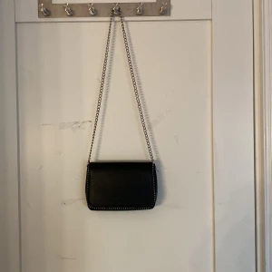 Svart axelväska med kedjerem - Stilren svart axelväska med silverfärgad kedjerem och dekorativa nitar längs kanterna. Väskan har en klassisk, rektangulär form och är perfekt för att bära det viktigaste. Enkel att matcha med olika outfits och ger en cool touch till din stil.