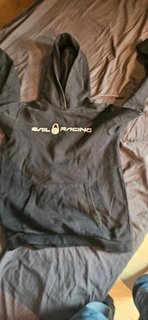 Svart hoodie från Sail Racing - Svart hoodie från Sail Racing med stor logga tryckt på bröstet. Klassisk huva och magficka framtill. Mjuk insida och ribbade muddar vid ärmslut och nederkant. Pris kan diskuteras