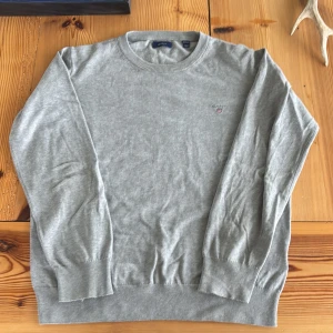 Grå bomullströja från GANT - Storlek 164 men passar upp emot 170.          Snygg grå tröja från GANT med rund hals och diskret logga broderad på bröstet. Tröjan är långärmad och har ribbade muddar vid ärmslut och nederkant. Perfekt för dig som gillar stilrena och klassiska plagg med en modern touch.  