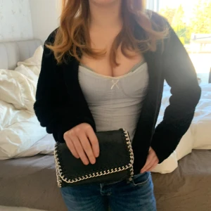 Clutch väska  - Snygg svart kuvertväska i skinnimitation med coola silverfärgade kedjedetaljer runt kanterna. Väskan har en stilren, rektangulär form och stängs med lock. Perfekt accessoar för att lyfta din outfit.