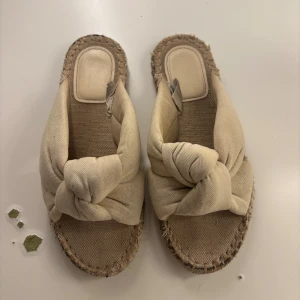 Beige sandaler med knut framtill - Säljer ett par beige sandaler med grov sula och snygg knutdetalj framtill. Ovandelen är i mjukt tyg och sulan har en rå, fransad kant som ger en chill vibe. Perfekta för varma dagar och enkel att slänga på sig.