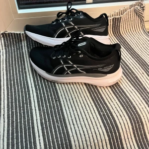 Svarta Asics Gel-Kayano 31 sneakers - Säljer ett par svarta Asics Gel-Kayano 31 sneakers med vita och grå detaljer. Skorna har snörning, platt sula och rund tå. Ovandelen är i syntetmaterial med Asics logga på sidan och PureGEL-dämpning i sulan för extra komfort. Perfekta för träning och löpning.