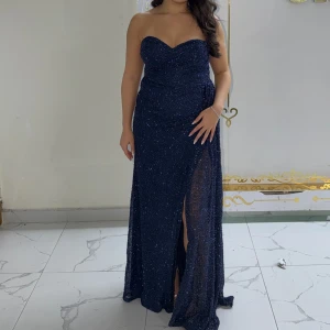 Marinblå glittrig långklänning - Säljer en marinblå glittrig långklänning med strapless design och en snygg slits framtill. Klänningen har en figurnära passform och skimrande material som ger en lyxig känsla. Perfekt för bal eller festliga tillfällen när du vill sticka ut. Klänningen hade passat för någon som har storlek 38 men på etiketten står det 42, den är liten i passform. Endast använd i några timmar. Priset kan diskuteras vid snabbaffär.