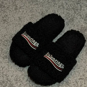 Svarta B innertofflor  - Mysiga svarta tofflor i storlek 41 med broderad logga och vågiga röda och vita linjer på ovansidan. Tillverkade i fluffigt syntetmaterial som ger en cool och unik look. Perfekt för chill dagar hemma eller när du vill sticka ut.