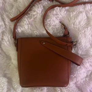 Brun axelväska i skinnimitation - Supersnygg brun axelväska med justerbar rem och dragkedja upptill. Väskan har en stilren, fyrkantig form och är tillverkad i skinnimitation. Axelrämen går att ta av om man vill. 