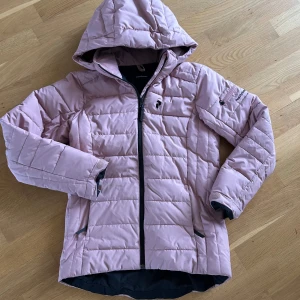 Gammelrosa skidjacka från Peak Performance - Säljer en gammelrosa skidjacka från Peak Performance med huva och svarta detaljer. Jackan har quiltad design, dragkedja framtill och två sidofickor. Perfekt för kyliga dagar och har en snygg, sportig look.