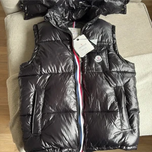 Svart dunväst från Moncler - Svart, glansig dunväst från Moncler med klassisk logotyp på bröstet. Västen har två sidofickor, dragkedja med röd, vit och blå kant, samt quiltad design. Materialet är polyester och dun, perfekt för lager-på-lager. Snygg detalj med serietidningsetikett på insidan. Skriv till mig för mer bilder, finns en huva som går att sätta på och ta av när som.