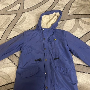 Blå parkajacka från Lyle & Scott - Blå parkajacka från Lyle & Scott med huva och mjukt vitt fleecefoder. Jackan har flera fickor med knappar, snörning vid midjan och klassisk gul logga på bröstet. Perfekt för kyliga dagar och har en stilren look som funkar till det mesta.