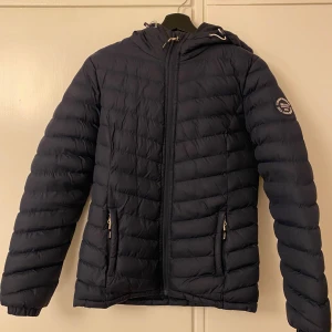 Mörkblå pufferjacka från Vinson Polo Club - Snygg mörkblå pufferjacka från Vinson Polo Club med huva och coolt badge på ärmen. Jackan har dragkedja framtill, två fickor med dragkedja och quiltad design. Perfekt för kalla dagar. 