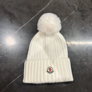 Vit ribbad mössa från Moncler - Vit ribbstickad mössa från Moncler med stor fluffig boll på toppen och uppvikt kant. Klassisk Moncler-logga framtill. Perfekt för kalla dagar och snygg till vinterlooken.