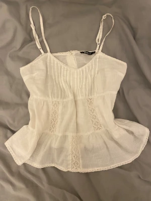 Vit linnetopp med spets från Gina Tricot - Supersöt vit linnetopp från Gina Tricot med tunna axelband och v-ringad hals! ❣️ Toppen har volang nedtill, ribbade detaljer framtill och inslag av spets i panelerna. Knappar bak i ryggen. 