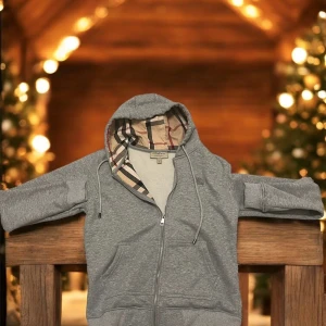 Grå hoodie från Burberry med dragkedja - Grå hoodie från Burberry med klassiskt rutigt foder i huvan och broderad logga på bröstet. Tröjan har dragkedja framtill, snörning i huvan och två fickor. Perfekt för dig som gillar stilren design med en touch av lyx.