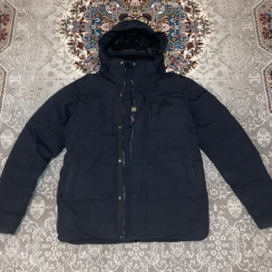 Svart pufferjacka från Five Seasons - Svart pufferjacka från Five Seasons med huva och dragkedja framtill. Jackan har en stilren look och är fodrad med mönstrat innerfoder. Perfekt för kalla dagar och har praktiska fickor på sidorna. Märkespatch på ärmen och insidan.