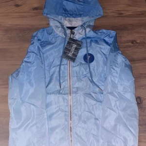 Blå vindjacka från Trapstar med huva - Snygg ljusblå vindjacka från Trapstar med huva och hel dragkedja framtill. Jackan har ett blått Trapstar-märke på bröstet och broderad logga på ryggen. Materialet är glansigt och lätt, perfekt för streetwear. Justerbara snören vid huvan och coola detaljer.