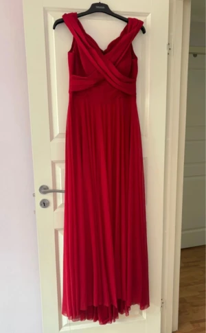 Röd långklänning med omlottband - Säljer en elegant röd långklänning med omlottband över axlarna och ett flytande, svepande tyg. Klänningen har breda axelband och ett vackert fall som ger en dramatisk look. Perfekt för dig som vill sticka ut med en klassisk och stilren siluett. Storlek 40🩷