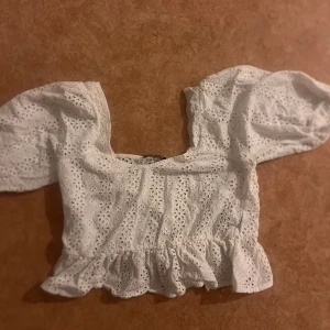 Vit blus med volang från Gina Tricot - Superfin vit blus från Gina Tricot i storlek 38. Blusen har broderade hålmönster, puffiga korta ärmar och en söt volangkant nertill. Perfekt för en somrig look med romantiska vibes och fyrkantig ringning.