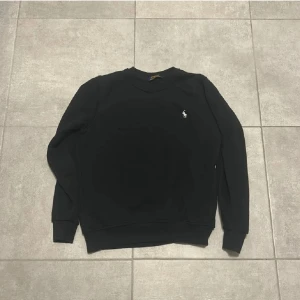 Svart sweatshirt från Polo Ralph Lauren - Svart sweatshirt från Polo Ralph Lauren med klassisk rund hals och broderad vit logga på bröstet. Tröjan har långa ärmar och ribbade muddar vid ärmslut och nederkant. Perfekt för en clean och stilren look.