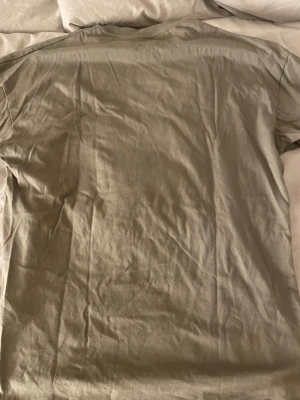 Beige Stüssy t-shirt med tryck - Säljer en beige t-shirt från Stüssy med klassiskt vitt logotryck på bröstet och texten 'new york london tokyo los angeles paris' under. T-shirten har rund hals och korta ärmar, tillverkad i mjuk bomull för skön känsla. Perfekt för dig som gillar streetwear.