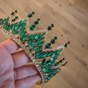 Glittrande grön krona tiara med kristaller - En riktigt maffig tiara i guldton med gnistrande gröna kristaller och klara stenar. Tiaran har spetsig form och är täckt av smaragdgröna stenar i olika former, vilket ger en lyxig och kunglig känsla. Perfekt för dig som vill sticka ut och glänsa på nästa stora event.Elegant krona med gröna och klara strasstenar. Perfekt för speciella tillfällen när du vill känna dig extra fin.