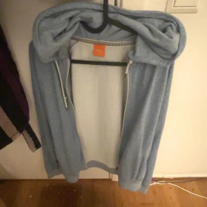 Ljusblå hoodie från Hugo Boss - Säljer en chill ljusblå hoodie från Hugo Boss med huva och snörning. Tröjan är i mjuk frotté och har ribbade muddar vid ärmslut och nederkant. Perfekt för att slänga på sig när man vill vara bekväm. Märke syns tydligt på orange etikett i nacken.