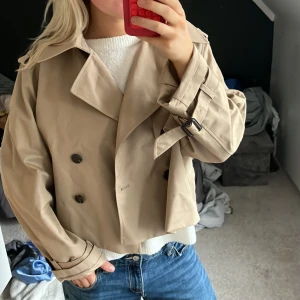 Beige croppad trenchcoat från H&M - Snygg croppad trenchcoat från H&M i beige med dubbelknäppning, stora slag och bälte vid ärmsluten. Jackan har en loose fit och är tillverkad i ett slätt polyestertyg. Perfekt för dig som vill ha en trendig och stilren look.