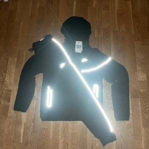 Svart Nike dress  med reflexdetaljer - Svart vindjacka från Nike med huva och vita reflexdetaljer längs bröstet, ärmarna och fickorna. Jackan har dragkedja framtill och två fickor med dragkedja. Perfekt för dig som vill sticka ut i mörkret och samtidigt ha en sportig look.