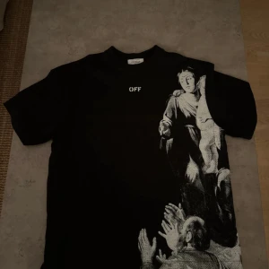 Svart Off-White t-shirt med print - Svart t-shirt från Off-White med vit text 'OFF' på bröstet och ett stort konstnärligt print i vitt på framsidan. Baksidan har Off-White-loggan i vitt. Klassisk rund hals och korta ärmar. Materialet är mjuk bomull som känns skön mot huden. Finns med prislapp. Äkta 