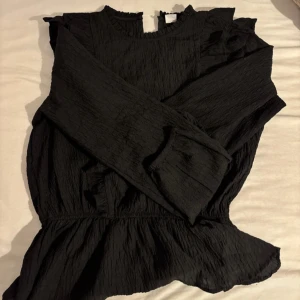 Svart blus med volangdetaljer - Säljer en svart blus med långärmade och volangdetaljer längs axlar och bröst. Blusen har en rundad hals och är tillverkad i ett lätt, krinklat tyg som ger en cool struktur. Midjan är markerad och nederdelen har en lätt peplum-effekt. Perfekt för dig som gillar stilrena och trendiga plagg.