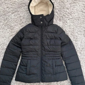 Svart pufferjacka med teddyfoder - Svart pufferjacka med mysigt vitt teddyfoder på insidan och huva. Jackan har dragkedja framtill, två fickor och snygga quiltade detaljer. Passformen är normal och jackan är perfekt för kalla dagar. Stängs med både dragkedja och tryckknappar vid halsen.