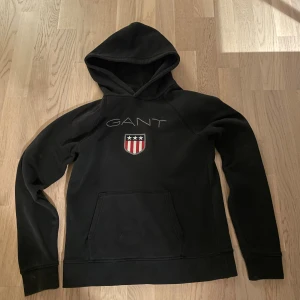  GANT hoodie  - Svart hoodie från GANT med huva och klassisk logga med sköld och stjärnor på bröstet. Tröjan har en magficka och långa ärmar. Perfekt för dig som gillar stilren design och vill ha en bekväm tröja med coolt tryck.