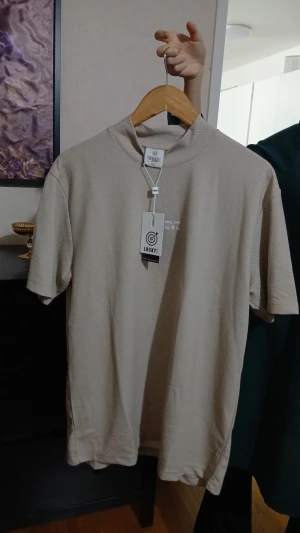 Beige t-shirt från Lucky med text - Snygg beige t-shirt från Lucky med diskret texten 'BELIEVE' på bröstet. T-shirten har en lite högre rund hals och korta ärmar. Materialet är mjukt och har en lätt strukturerad yta som ger en cool känsla. Köpt från Turkiet aldrig använt eftersom den va för stor. Klicka på bilden