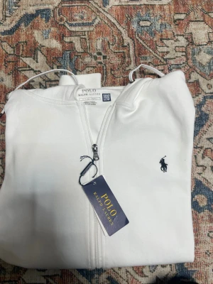 Polo Ralph Lauren - Vit hoodie från Polo Ralph Lauren med klassisk broderad logga på bröstet. Tröjan har huva med snörning, dragkedja framtill och två fickor. Tillverkad i mjuk bomull för skön komfort. Perfekt för chill dagar och enkel att matcha med jeans eller joggers.