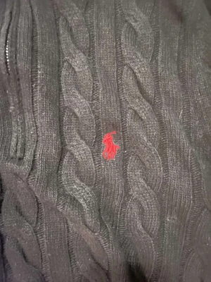 Svart stickad half zip tröja Polo Ralph Lauren - Svart kabelstickad tröja från Polo Ralph Lauren med röd broderad logga på bröstet. Tröjan har ribbad krage, half zip-dragkedja och långa ärmar. Materialet är mjuk ull och passformen är normal. Perfekt för dig som gillar klassisk stil med en modern touch.