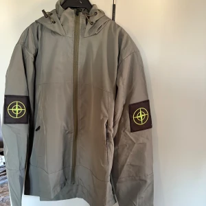  Vindjacka med huva - Snygg vindjacka  i beige med ikoniska patchar på båda ärmarna. Jackan har huva med snörning, dragkedja framtill och två sidofickor. Perfekt för dig som vill ha en clean och stilren look med street-vibe.