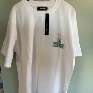 Vit Amiri t-shirt med fågeltryck - Vit t-shirt från Amiri med coolt tryck av två hägrar och vattenväxter på ryggen samt mindre tryck fram. Klassisk passform med rund hals och korta ärmar. Perfekt för dig som gillar streetwear med unik design.