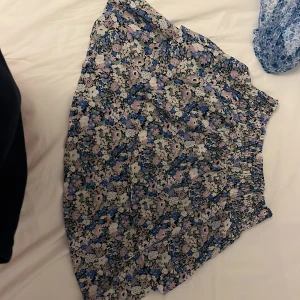 Blommig kortkjol från ONLY - Supersöt kortkjol från ONLY med blommigt mönster i blått, lila, vitt och svart. Kjolen har resår i midjan och är lätt och flowy, perfekt för varma dagar. Materialet känns mjukt och luftigt, och färgerna poppar verkligen. Perfekt att styla med en enkel topp.