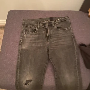 Grå jeans från H&M, storlek 32/32 - Säljer ett par grå jeans från H&M i storlek 32/32. Jeansen har en snygg tvättad look med slitningar och hål på ena benet. Klassisk femficksmodell och raka ben. Perfekta för en avslappnad streetstil.