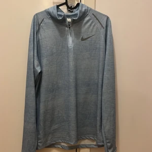 Nike ljusblå långärmad half zip tröja - Nike långärmad tröja i ljusblått med subtilt mönster och grå logga på bröstet. Tröjan har half zip framtill och är gjord i mjukt syntetmaterial som andas. Perfekt för träning eller chill, med sportig look och bekväm passform.