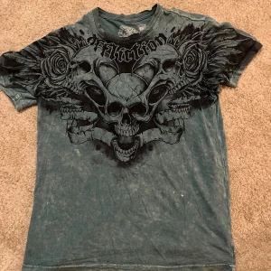 Blå Affliction t-shirt med döskalletryck - Säljer en blå t-shirt från Affliction med stort, detaljerat tryck av döskallar och rosor på framsidan. T-shirten har korta ärmar, rund hals och är tillverkad i 100% bomull. Bak finns ett litet svart tryck med korslagda spikar. Perfekt för dig som gillar edgy stil. Materialet är helt sjukt har aldrig sett en tröja med såhär bra material. 