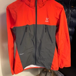 Röd och grå vindjacka från Haglöfs - Snygg vindjacka (gortex) från Haglöfs i rött/oranget och mörkgrått med huva och hel dragkedja. Jackan har två snedställda fickor med dragkedja och coolt mönstrat innerfoder. Perfekt för dig som gillar att vara ute och vill ha en sportig look. Jackan är i storlek 164 men passar även xs.