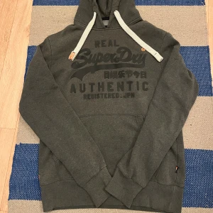 Grå Superdry hoodie med tryck - Snygg grå hoodie från Superdry med stort vitt tryck på bröstet och coola japanska tecken. Tröjan har huva med breda vita snören och ribbade muddar. Superdry orange label, perfekt för dig som gillar streetwear och vill sticka ut. Riktigt fint skick och är storlek Medium! Hör av dig vid några frågor!