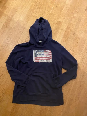 Mörkblå hoodie från Ralph Lauren - Snygg mörkblå hoodie från Ralph Lauren Denim & Supply med stor amerikansk flagga-patch och broderade detaljer på bröstet. Tröjan har huva, magficka och är tillverkad i 100% bomull. Perfekt för dig som gillar ”archive” stilen!! Storleken är M.
