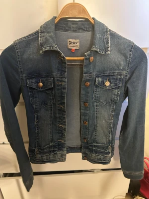 Blå jeansjacka från ONLY, strl 34 - Snygg klassisk jeansjacka från ONLY i blå denim. Jackan har långa ärmar, två bröstfickor med knapp och metallknappar framtill. Perfekt för dig som gillar en tidlös och chill stil. Passar dig som vill ha en jacka med normal passform och coola detaljer.