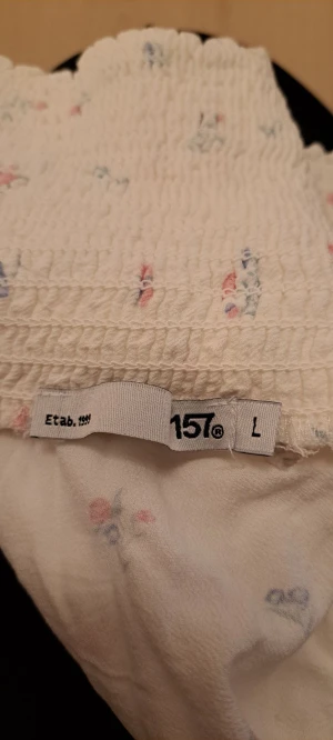 Vit blommig volangkjol från 157 - Supersöt kort kjol i vit med små blommor i rosa och blå toner från 157. Kjolen har smock upptill och volang nedtill som ger en skön och flowy känsla. Perfekt för dig som gillar romantisk och feminin stil. Materialet känns mjukt och lätt.