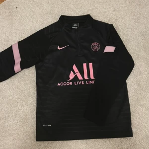 PSG fotbollströja Nike - Svart långärmad PSG träningströja från Nike med rosa detaljer och logga på bröstet. Tillverkad i lätt funktionsmaterial som andas.