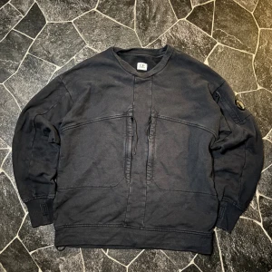 C.P. Company Sweatshirt  - Svart sweatshirt från C.P. Company med coola snörningsdetaljer framtill och den ikoniska linsen på ärmen. Tröjan har rund hals, långa ärmar och en loose passform. Perfekt för dig som gillar streetwear och vill ha något som sticker ut.