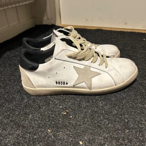 Golden Goose vita sneakers med stjärna - Golden Goose sneakers i vitt skinn med beige mockadetaljer och ikonisk stjärna på sidan. Skorna har svarta hälpartier, beige snörning och platt sula. Perfekta för dig som gillar en avslappnad och trendig look.